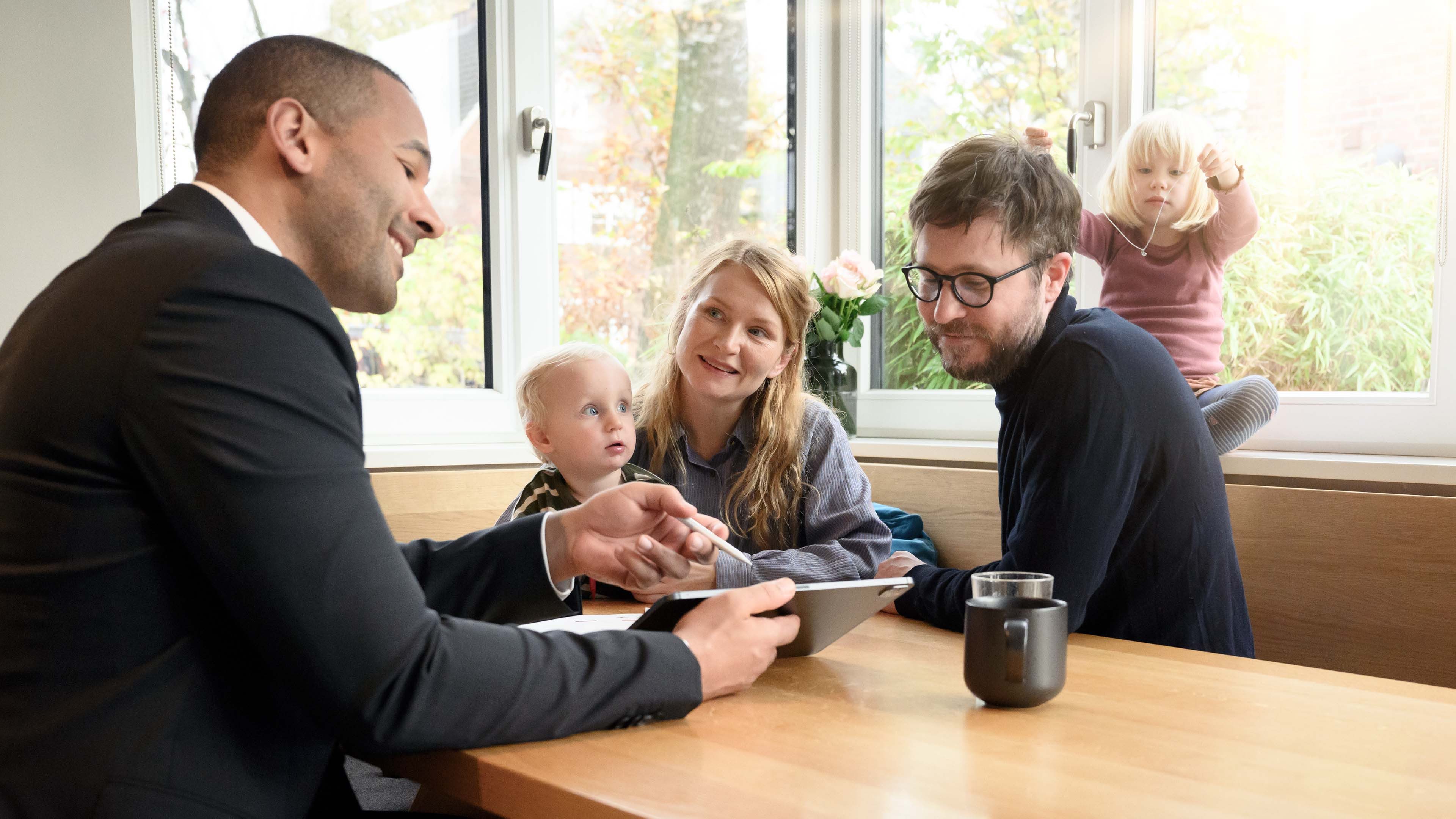 Ein Berater sitzt mit einer Familie an einem Holztisch und führt ein Gespräch, während er ein Tablet in den Händen hält. Die Familie sitzt einander zugewandt, und zwei Kinder sind ebenfalls anwesend, eines davon spielt auf der Fensterbank im Hintergrund.