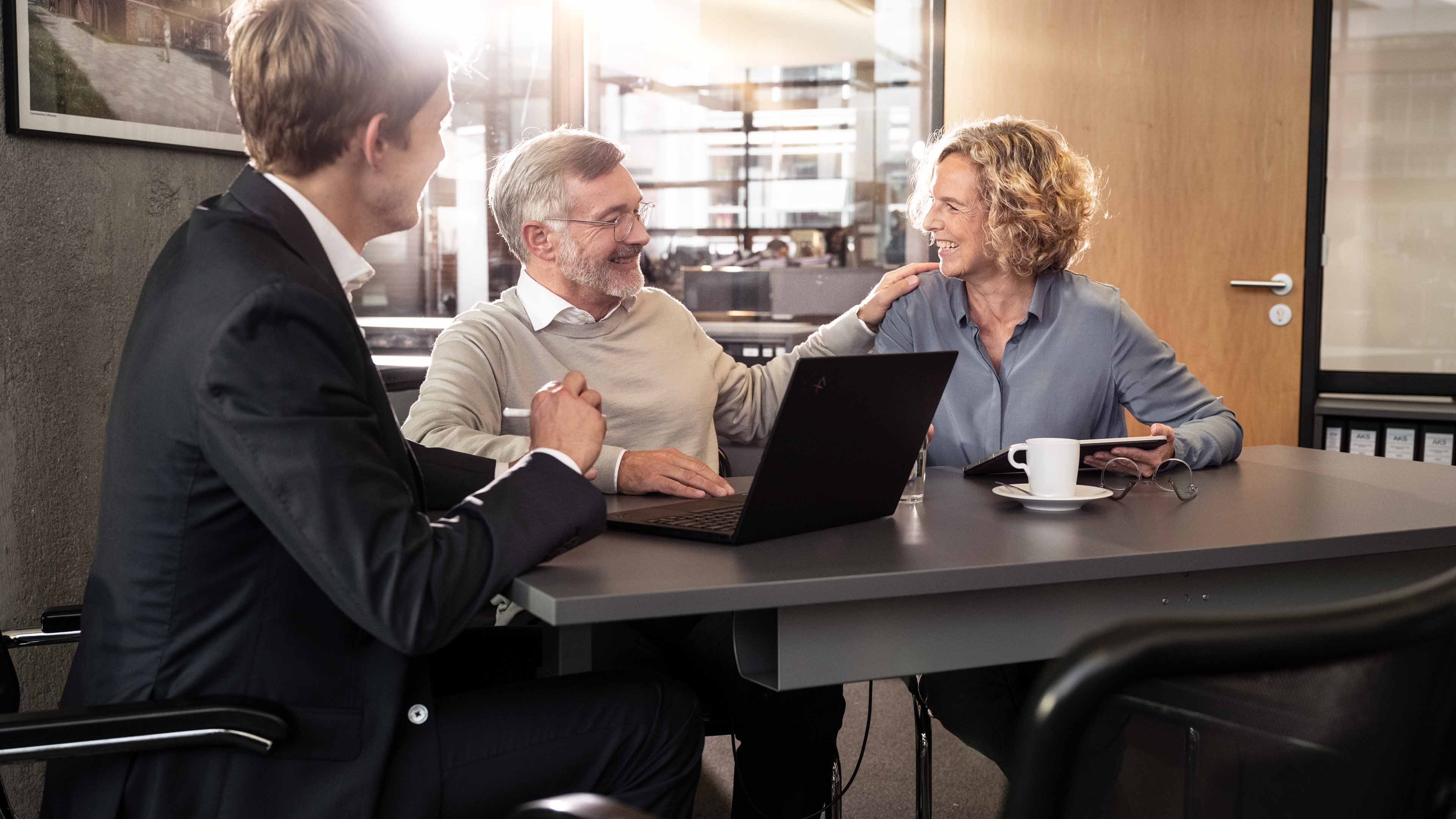 rei Personen sitzen gemeinsam an einem Tisch in einem modernen Büro und führen ein Gespräch, während ein Laptop, eine Tasse und ein Tablet vor ihnen liegen. Eine Person gestikuliert, während die anderen aufmerksam zuhören und miteinander interagieren.
